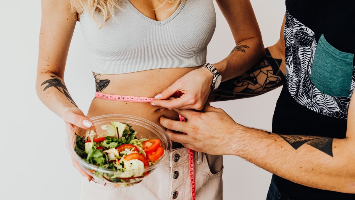 A couple measuring waistline with a tape and holding a fresh salad bowl, focusing on fitness and weight loss.